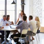 Businesswoman presenting to colleagues at a meeting about nonprofit jobs in a conference room at a table with six other people in a sunny room