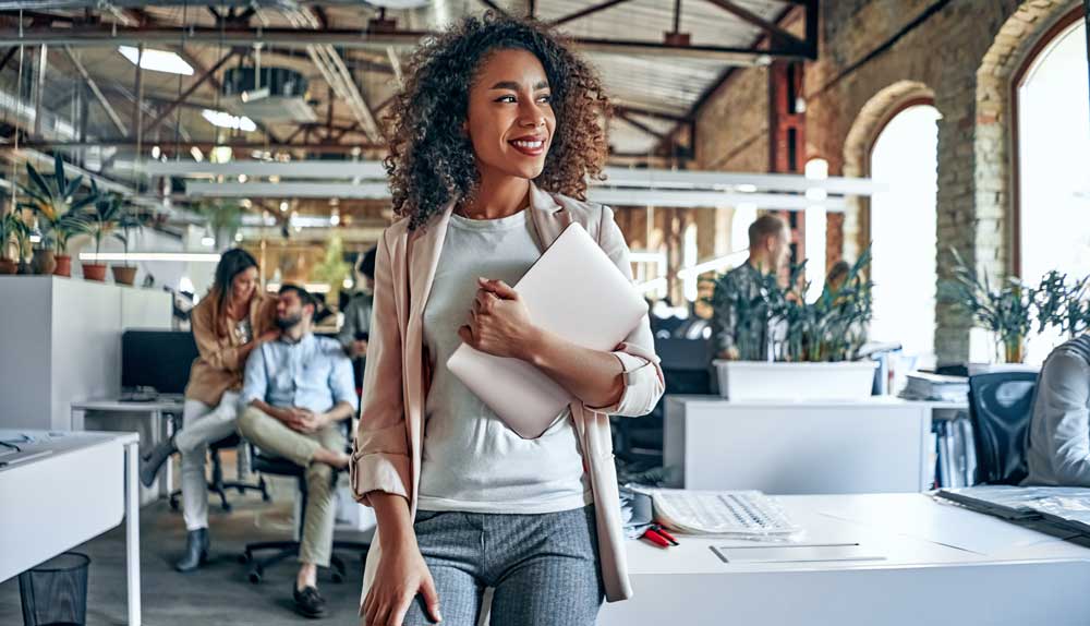 A women newly hired at a nonprofit looks around her new office after finding a new nonprofit job. She is standing and smiling with a laptop in her hand.