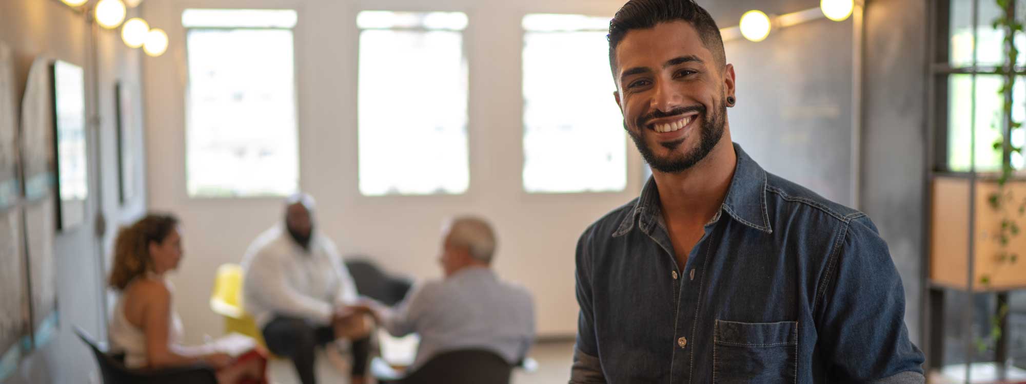 Nonprofit Jobs - Foundation List Nonprofit job board postings - man smiling at you dressed in a denim shirt. In the background a nonprofit meeting of three people can be seen.