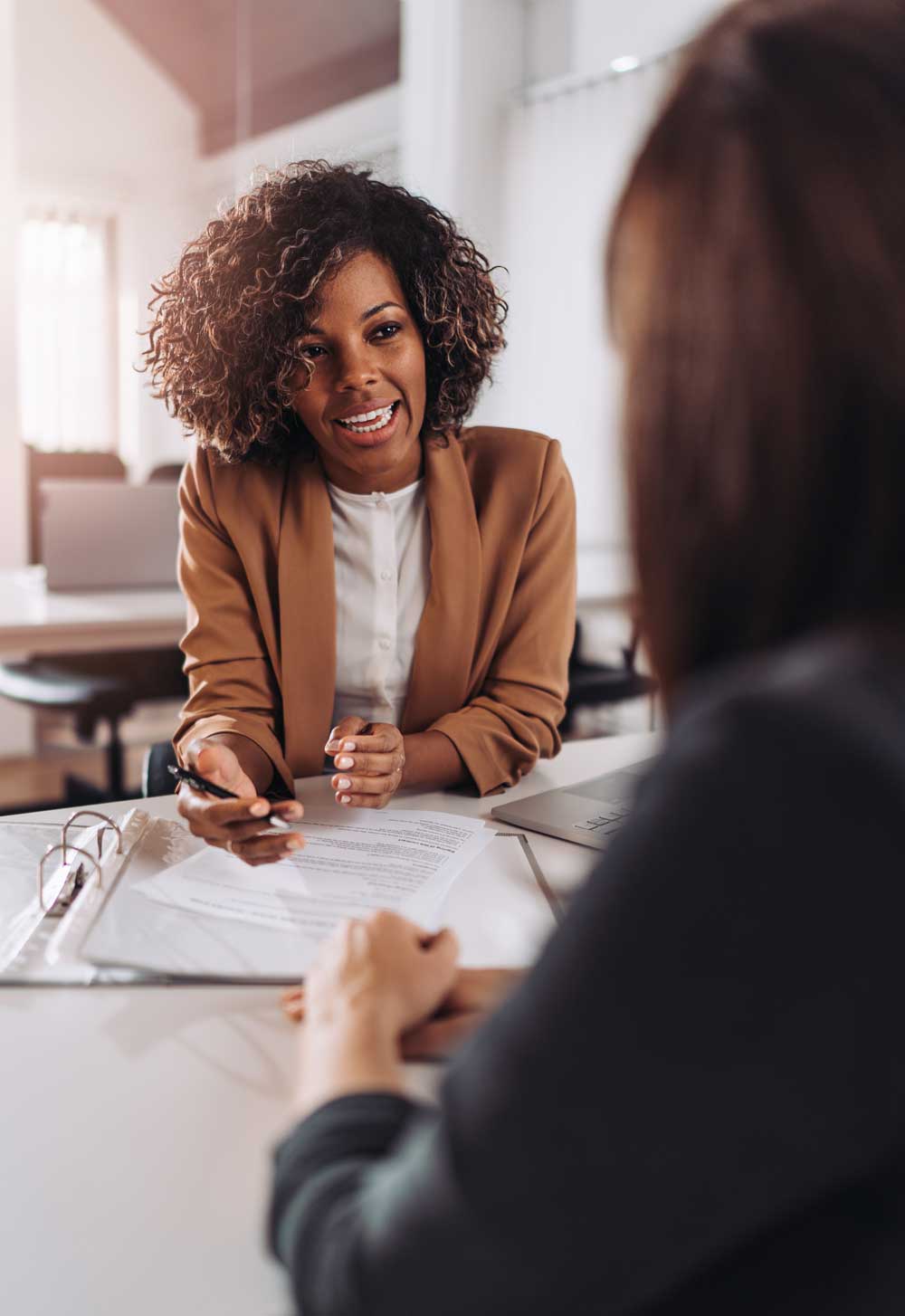 A women interviews a nonprofit job candidate for a foundation job. She is holding a pen and speaking to the candidates. Two people are seen seated at an interview. A resume can be seen on the table.