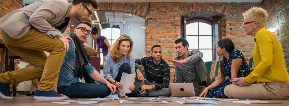 A team at a great nonprofit looks over resumes for job candidates. Resumes can be seen on the floor. Seven people are seated and discussing nonprofit employment dress professionally and in a open office.