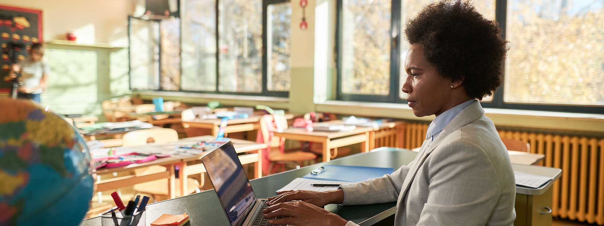 An elementary school teacher works at her computer in her empty classroom, looking at educational job opportunities